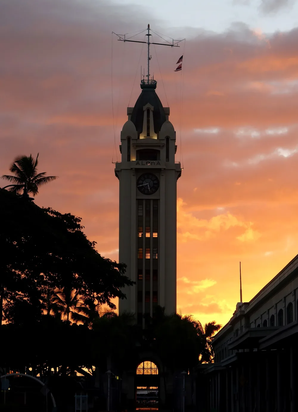 Aloha Tower silhouetted against an orange and pink sunset sky above palm trees